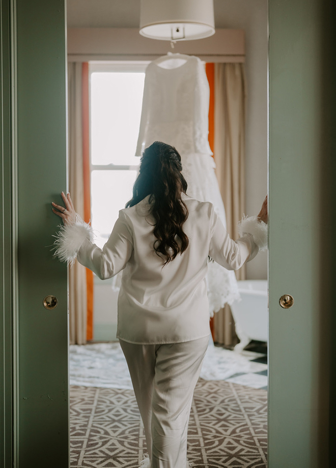 Bride wearing white satin pajamas walking into the suite at Hotel ICON with her dress hanging from the chandelier. 