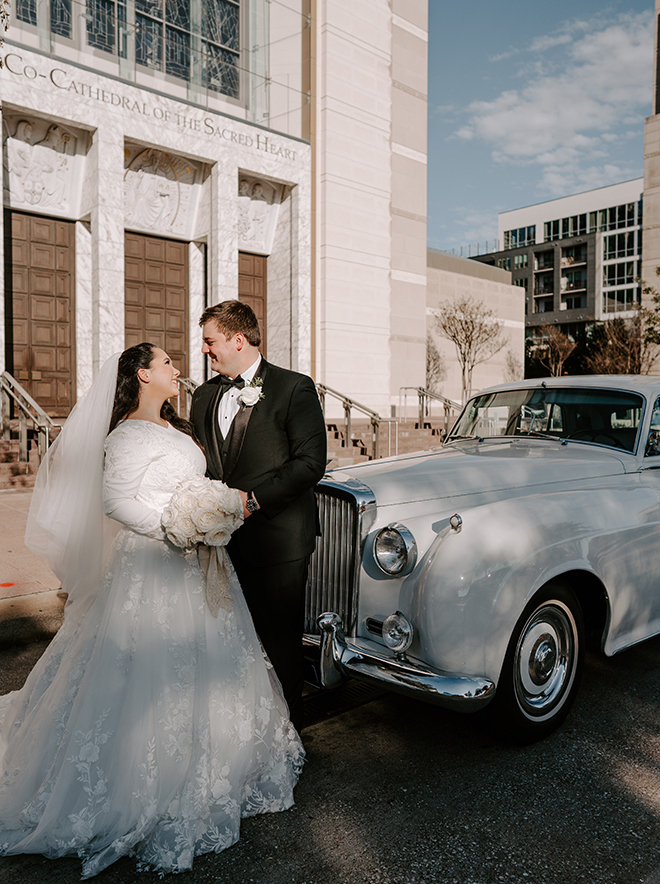 Bride and groom smiling at each other in front of the church and a white vintage Bentley. 