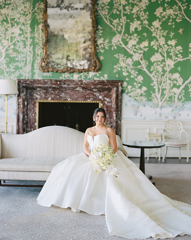 A bride sitting on a couch in a room with green and white floral wallpaper. 