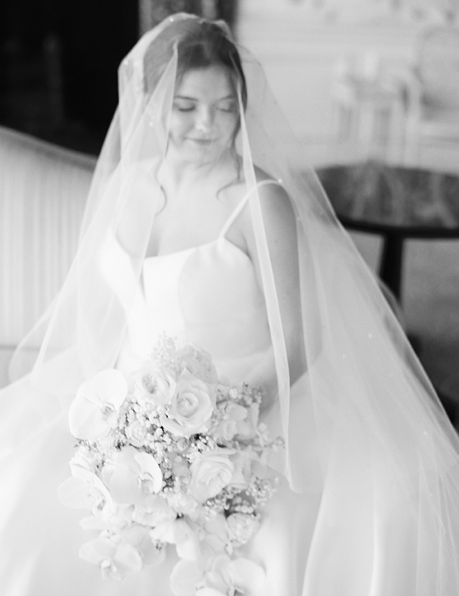 A black and white photo of the bride sitting down holding a bouquet with a veil over her head. 