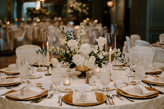 A round reception table with gold plates, white floral centerpieces and candles. 