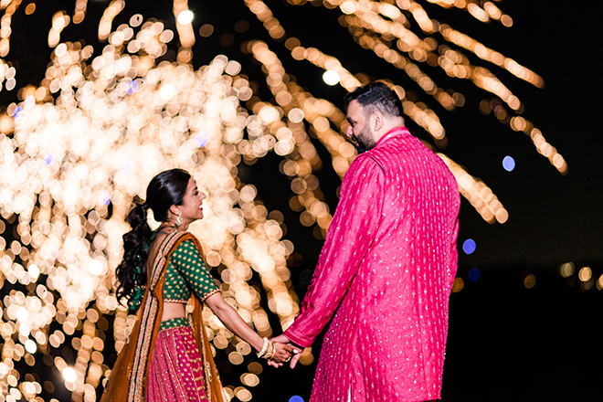 A bride and groom holding hands and smiling at each other during a fireworks show. 