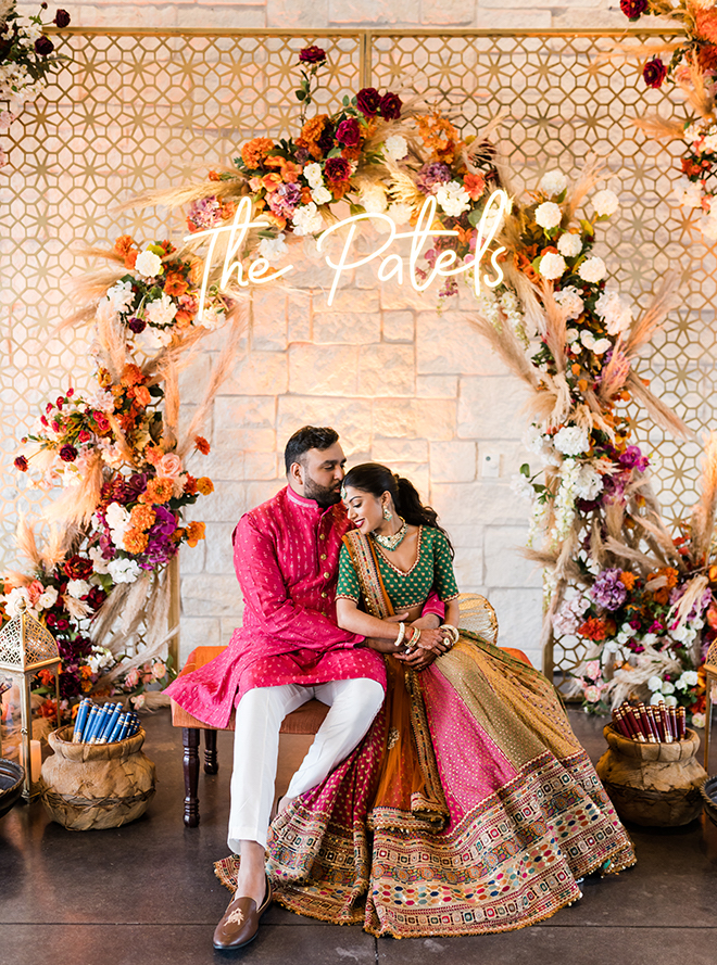 Bride and groom sitting on a bench under a floral arch with a sign that says "The Patels."