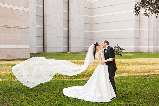 The bride's tule veil flows far in the wind as the broom holds her close.