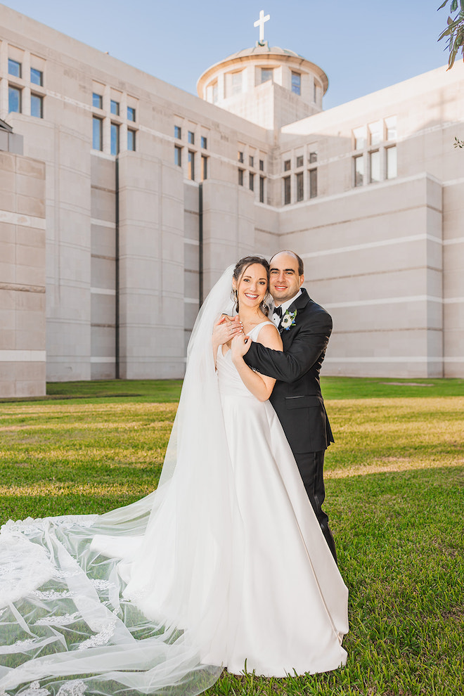 Outside of the church, the bride and groom pose closely for photos in their suit and gown. 