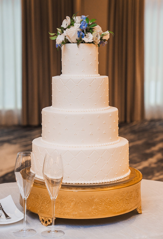 The delicious cake stands tall with 4 tiers, white icing, and white and blue florals on top. 