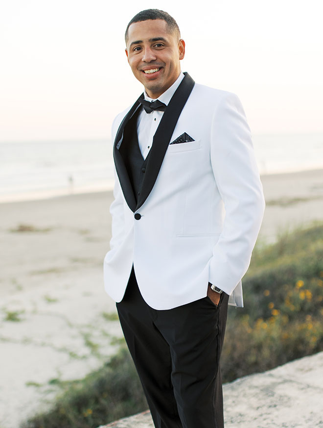 The groom wearing a white tuxedo stands along the Galveston seawall. 