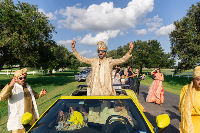 The groom standing up in a convertible yellow car for his Baraat. 